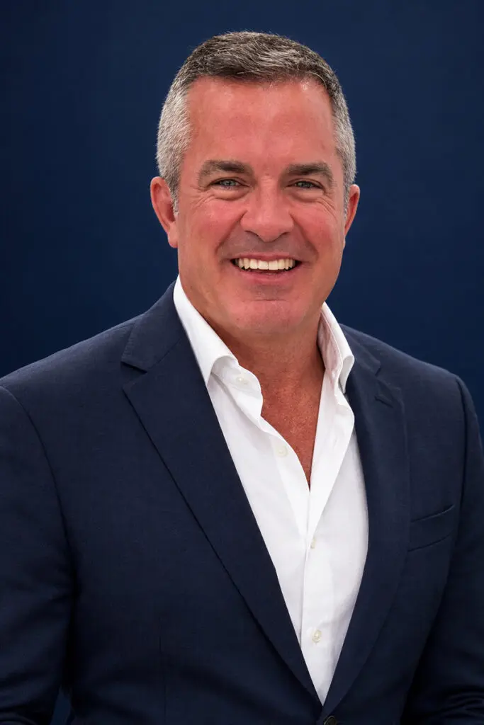 Man with short gray hair smiling, wearing a navy suit and white shirt.