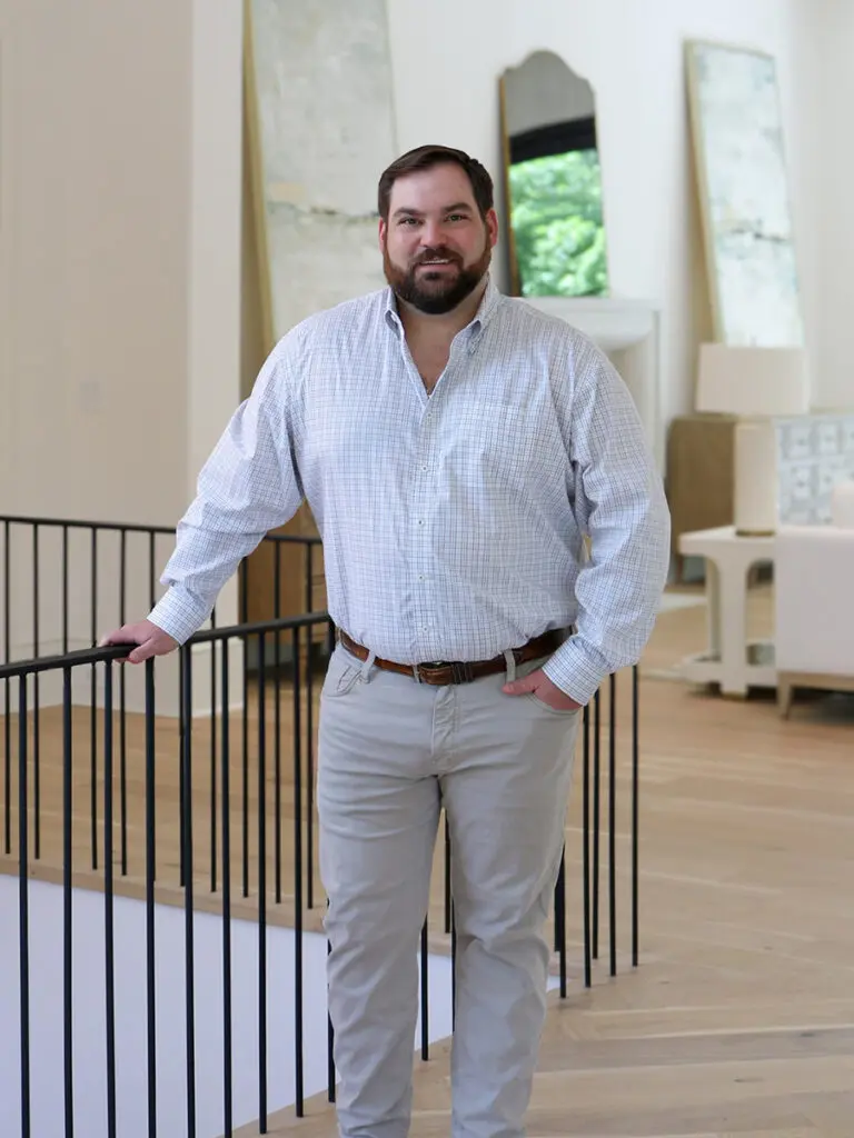 Man in light blue plaid shirt and gray pants standing near a staircase railing.